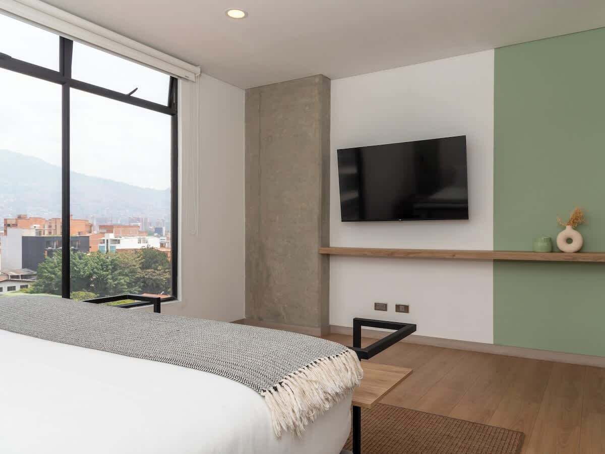A modern hotel room with a large bed, a flat-screen TV mounted on the wall, and a view of the city through large windows.