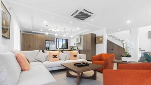 A modern living room with a sofa, armchairs, and a coffee table. There is a kitchen in the background with wooden cabinets and a staircase. - Stylish 3BR Villa by Vistacana Rentals (Punta Cana)
