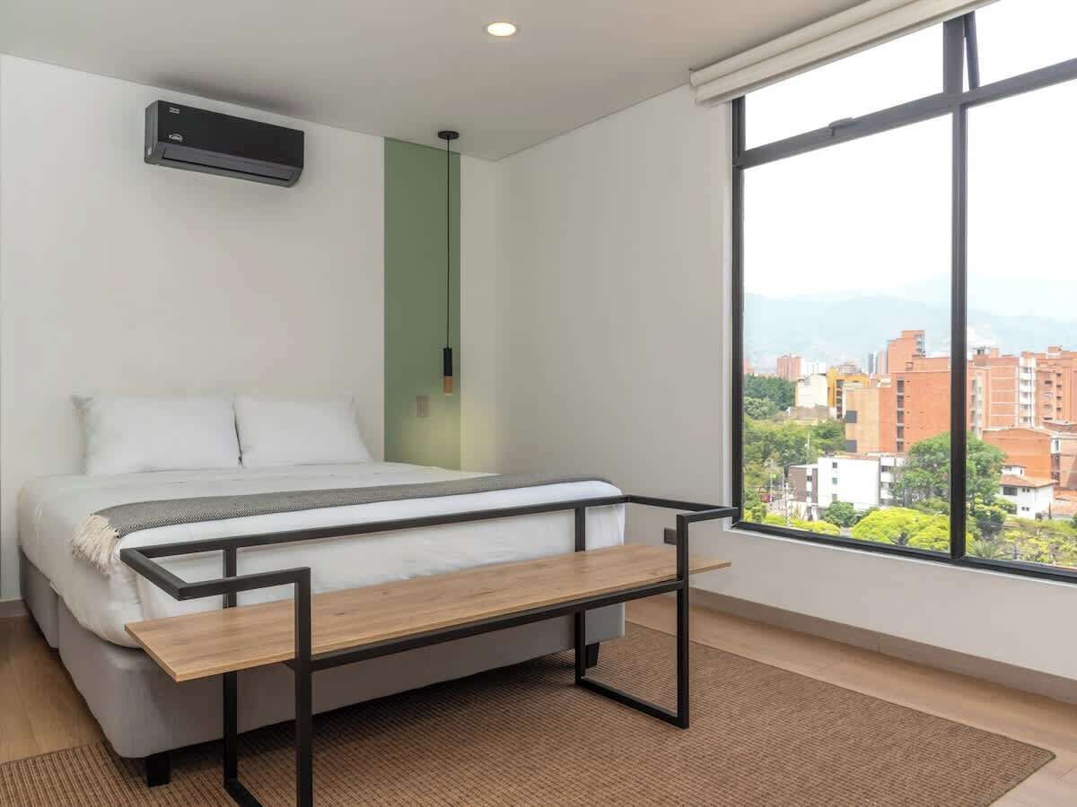 A bedroom with a bed, a bedside table, a window with a city view, and an air conditioning unit on the wall.