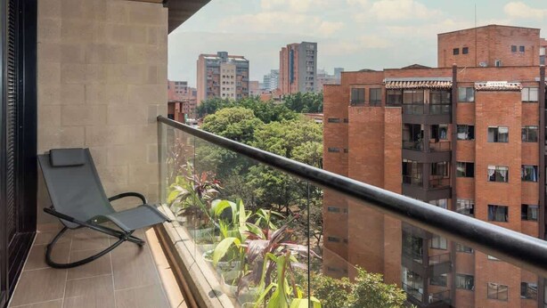 Ein Balkon mit Stuhl, Glasgeländer und Blick auf Stadtgebäude und Bäume.