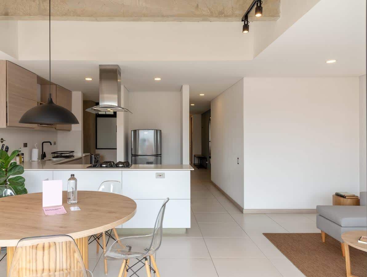 A modern kitchen with a central island, white cabinetry, and stainless steel appliances. There is a dining area with a round table and chairs.