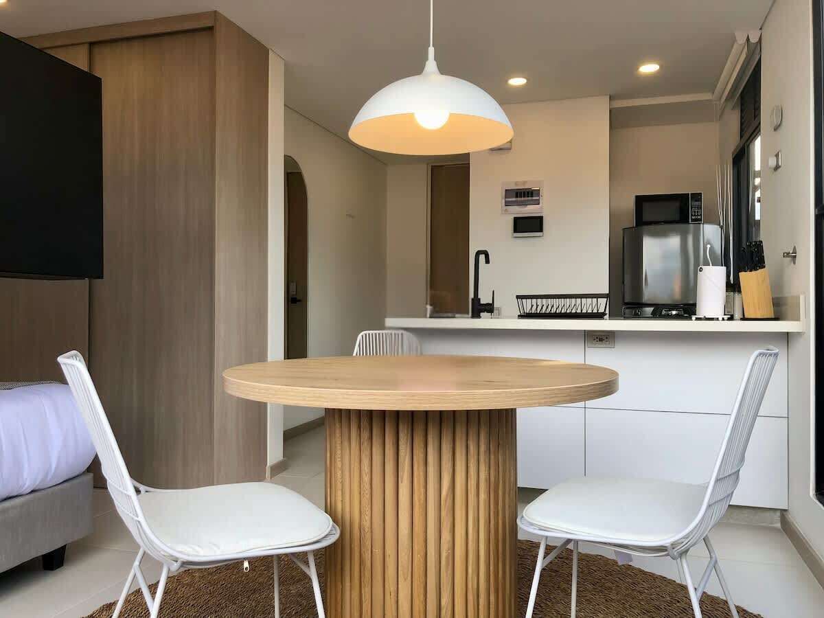 A modern kitchen with a round wooden table, white chairs, a hanging light fixture, and a built-in microwave.