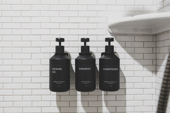 Drei schwarze Flaschen mit Duschgel, Shampoo und Conditioner auf einer weißen Fliesenwand.