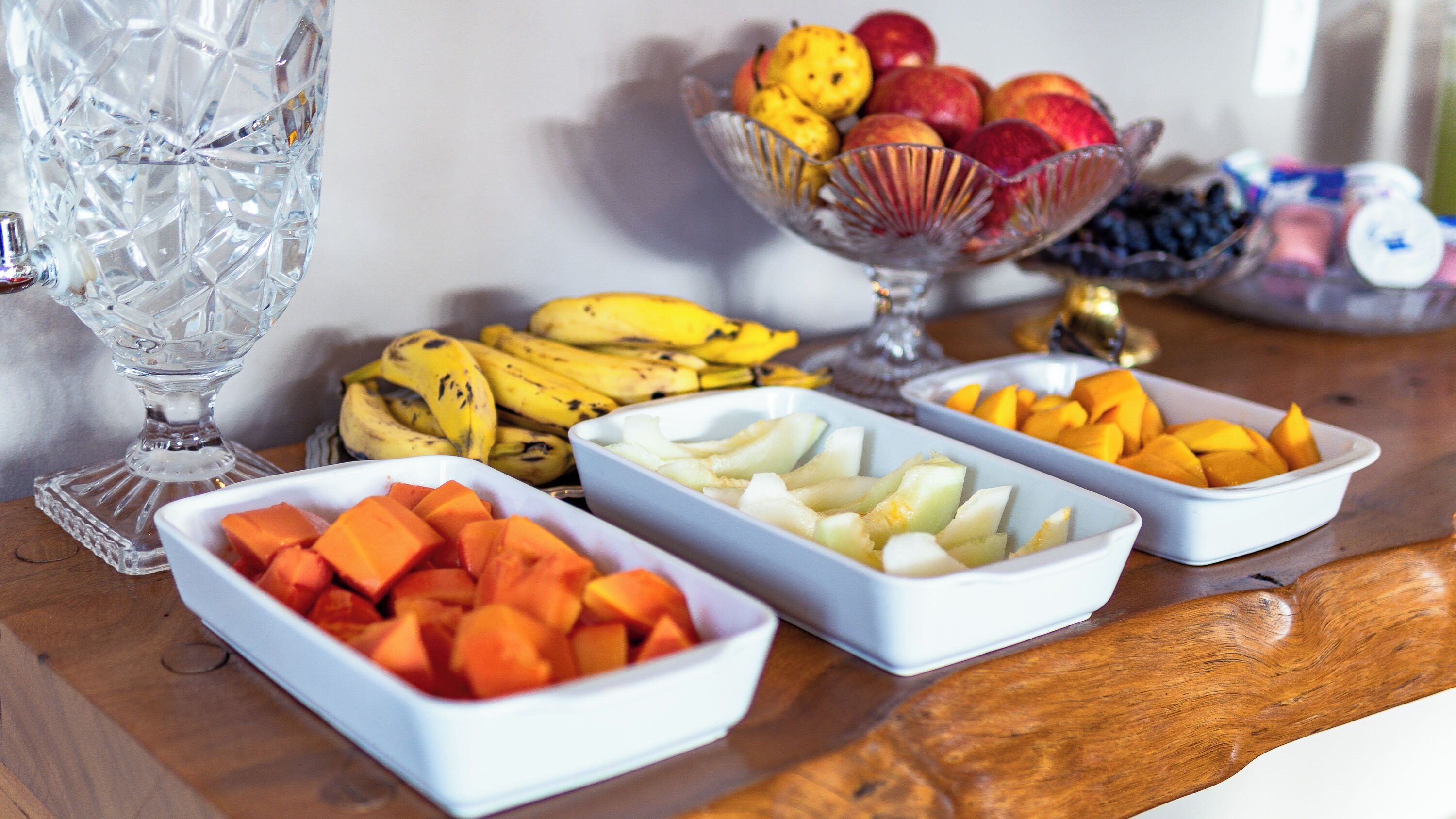 A table with various fruits in white bowls, a glass vase, and a crystal decanter.