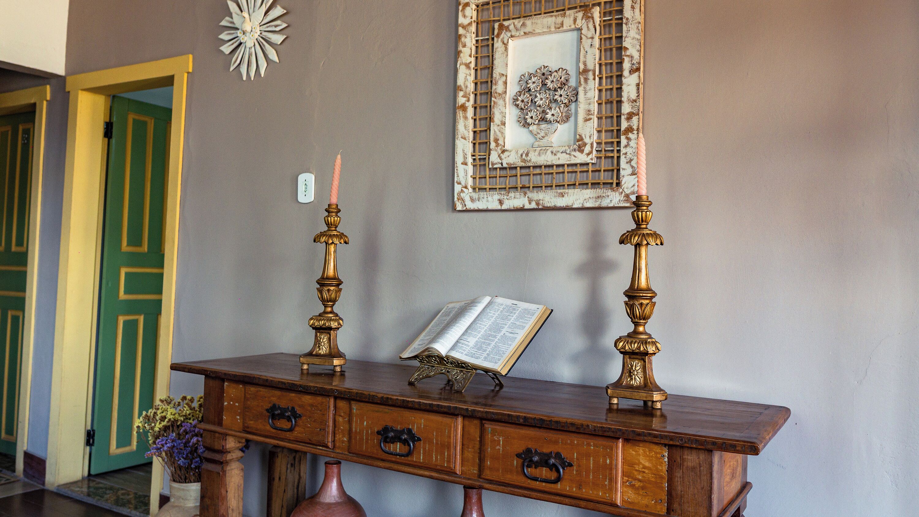 Una consola de madera con un libro, dos candelabros y un florero con flores.
