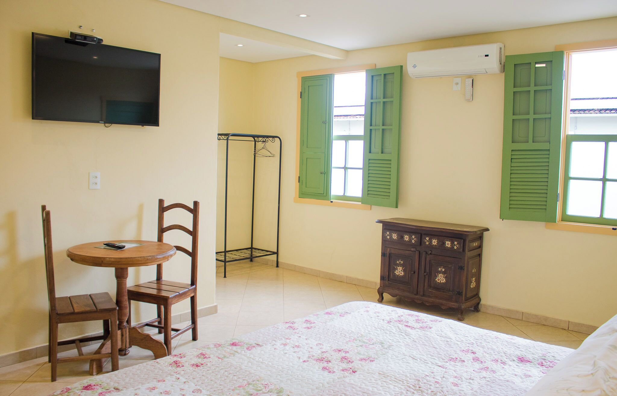 Une chambre à coucher comprenant un lit, une chaise en bois, une petite table, une armoire, un téléviseur fixé au mur et une fenêtre avec des volets verts.