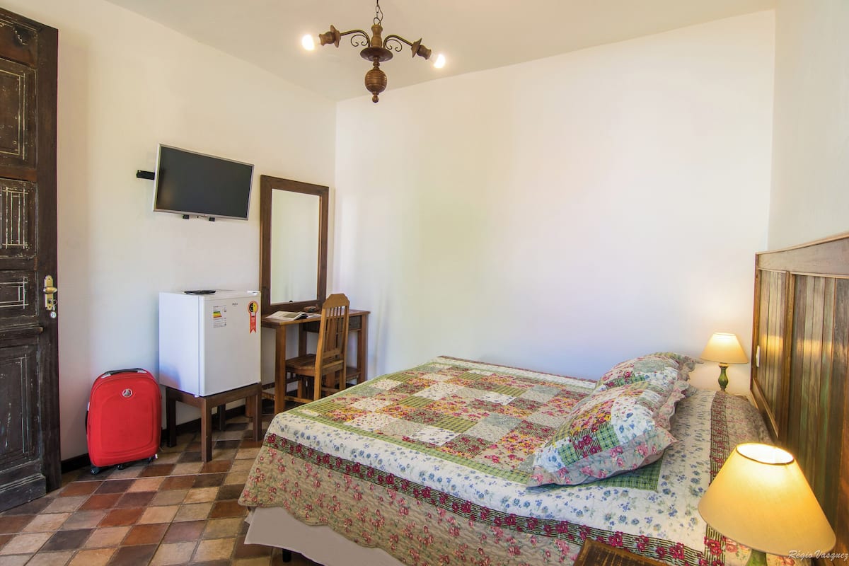 Ein Zimmer mit einem Bett, einem Schreibtisch, einem Sessel, einem Fernseher und einem roten Koffer.
