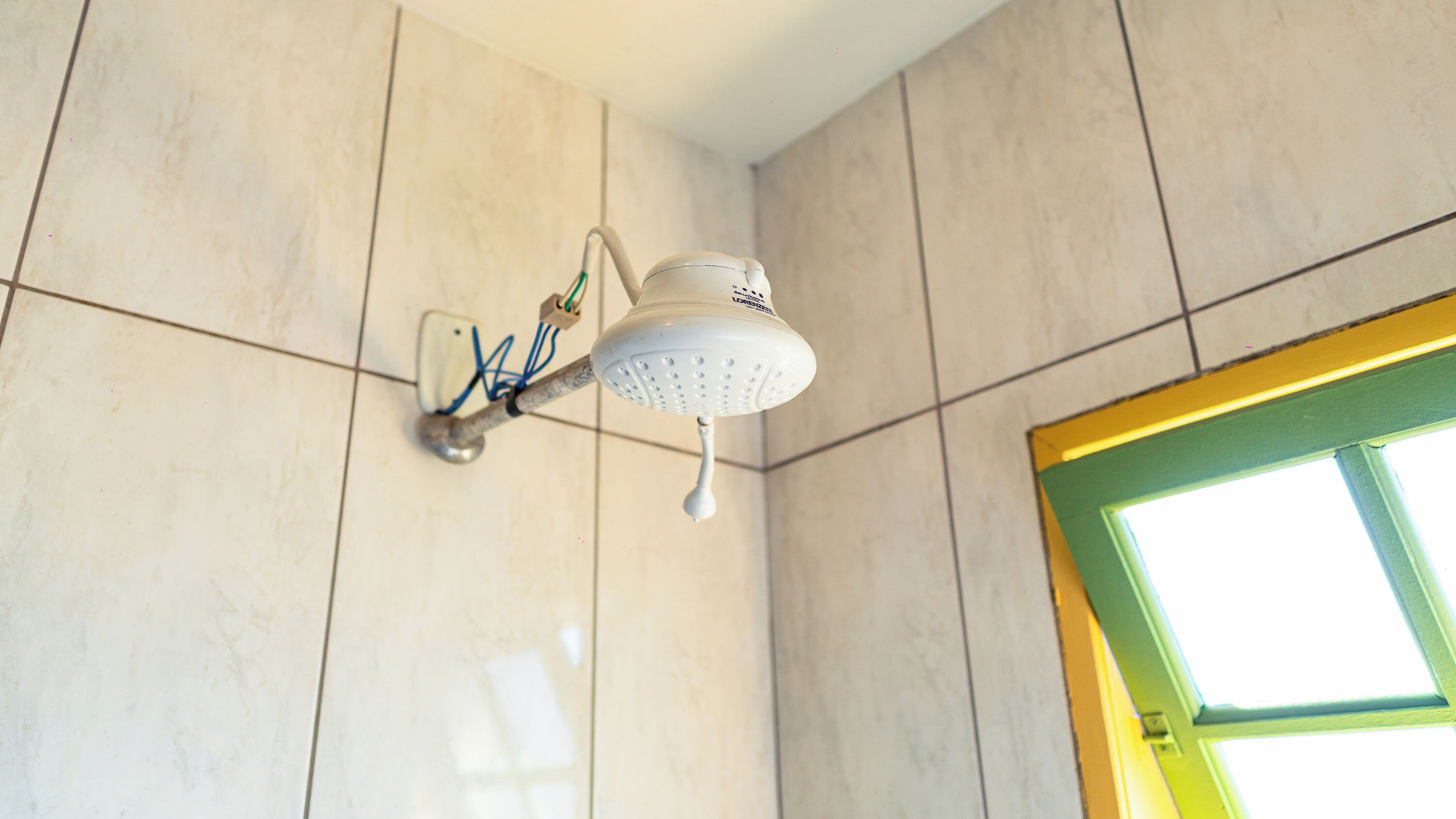 Un cabezal de ducha instalado en una pared con azulejos, con un enchufe eléctrico a la vista y una ventana con marco verde.