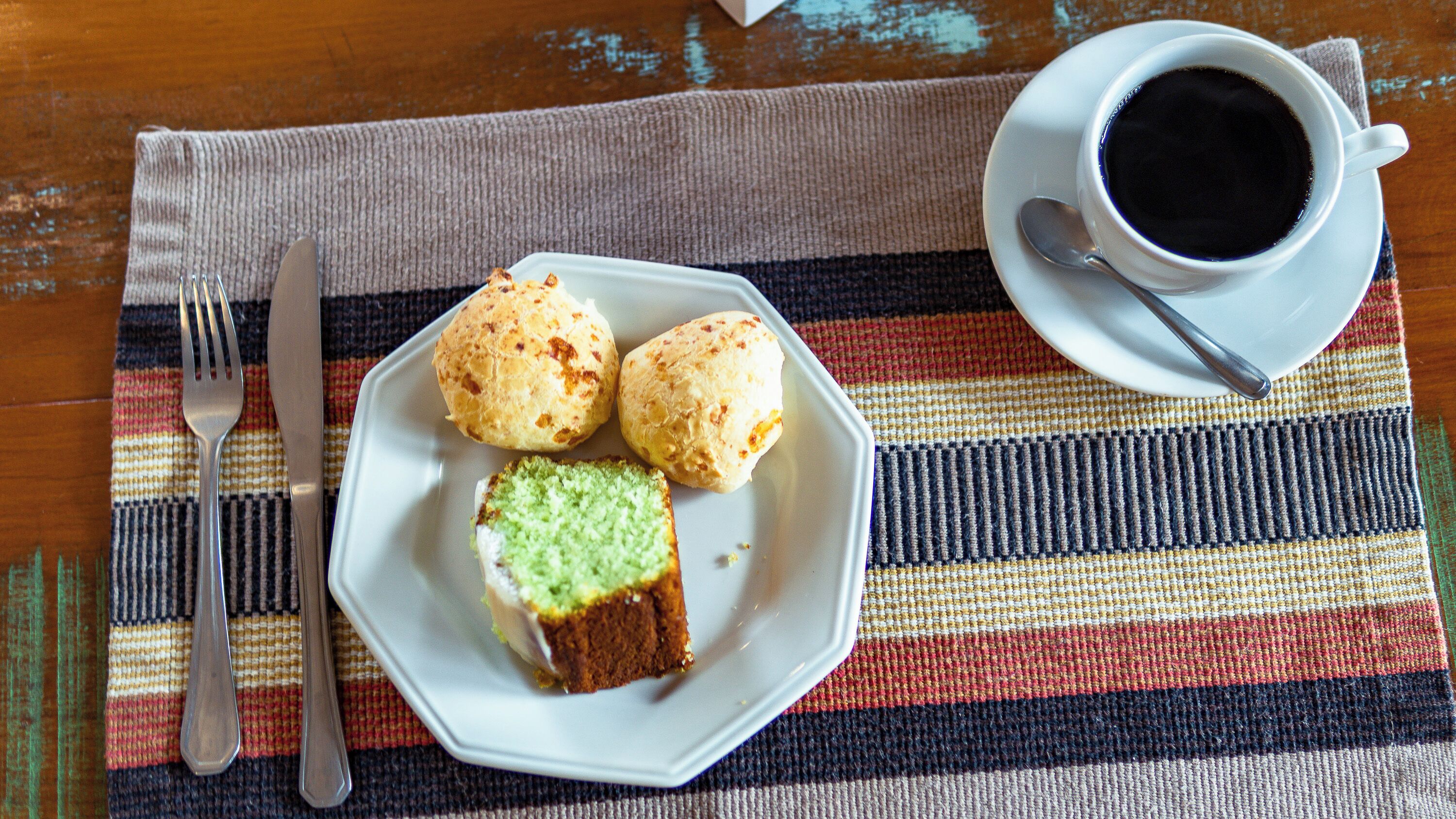 A plate with two pastries and a slice of cake on a striped placemat, a cup of coffee, and cutlery on a wooden table.