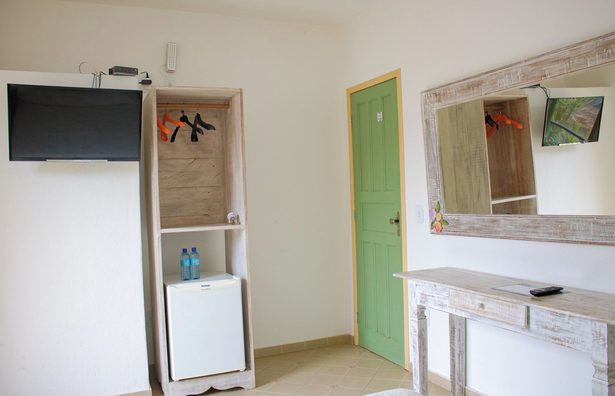 A room with a television mounted on the wall, a small white refrigerator, a wooden cabinet with hanging items, a green door, and a mirror with a decorative frame.