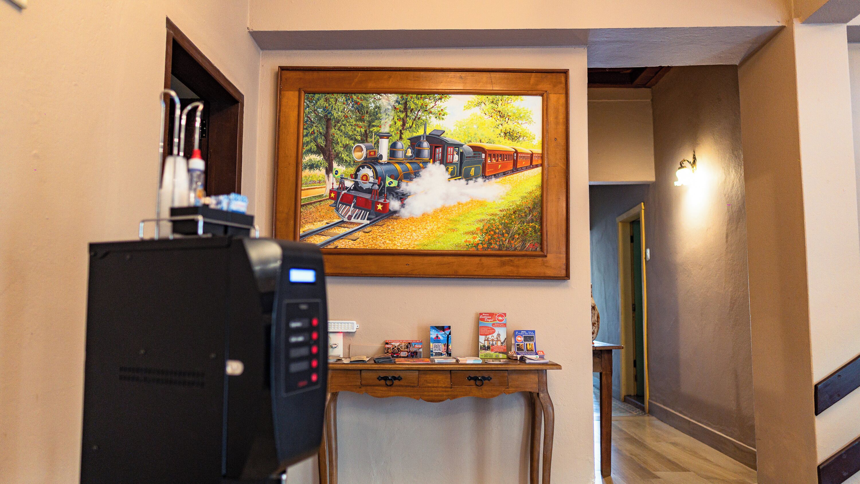 Un vestíbulo de hotel con un cuadro de un tren, una máquina de café, y una pequeña mesa con libros y un teléfono.