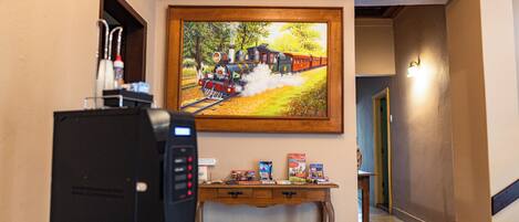 Un vestíbulo de hotel con un cuadro de un tren, una máquina de café, y una pequeña mesa con libros y un teléfono.