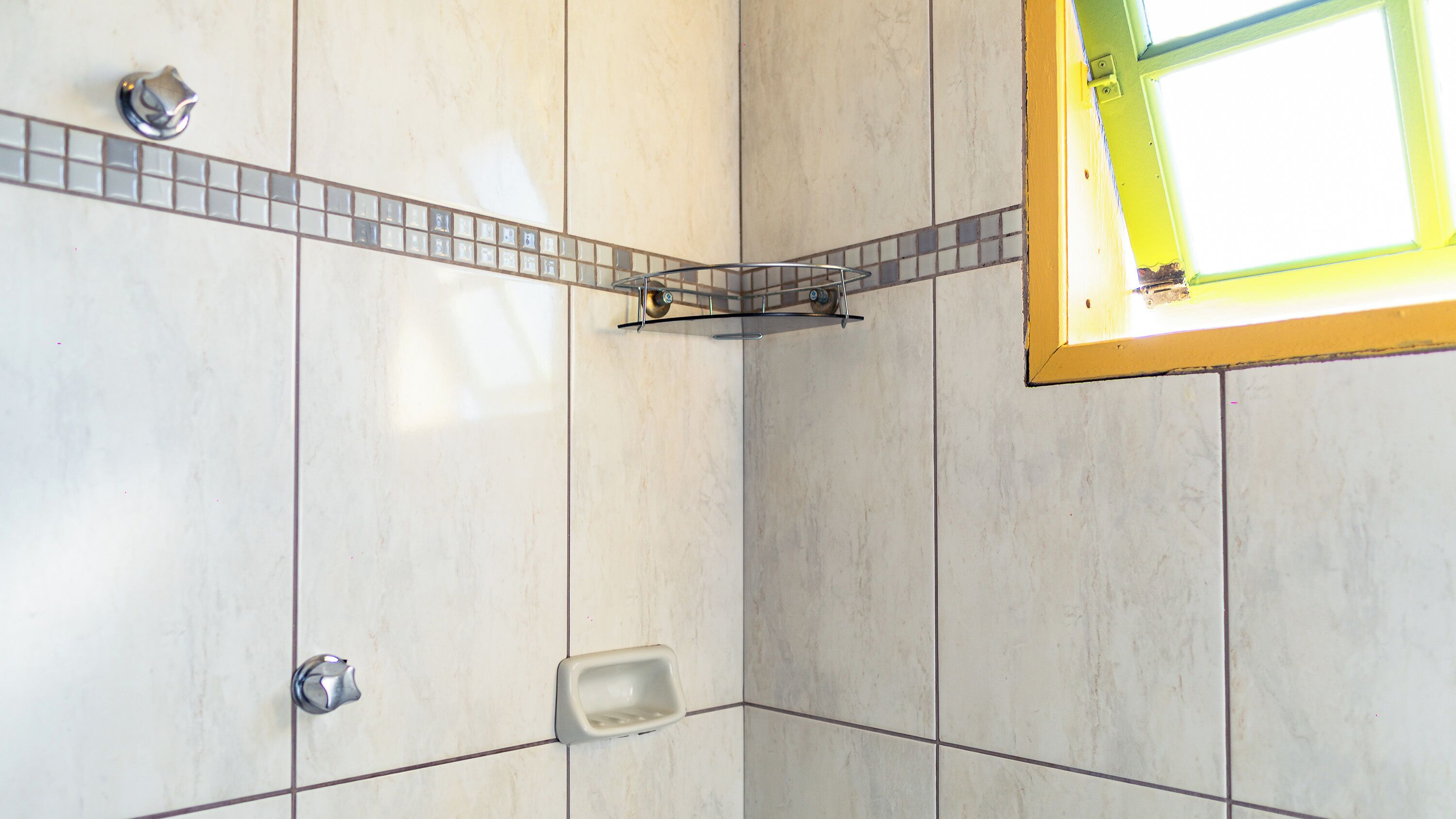 Une salle de bain avec un sol carrelé, un espace douche et une fenêtre dotée d’un cadre jaune.