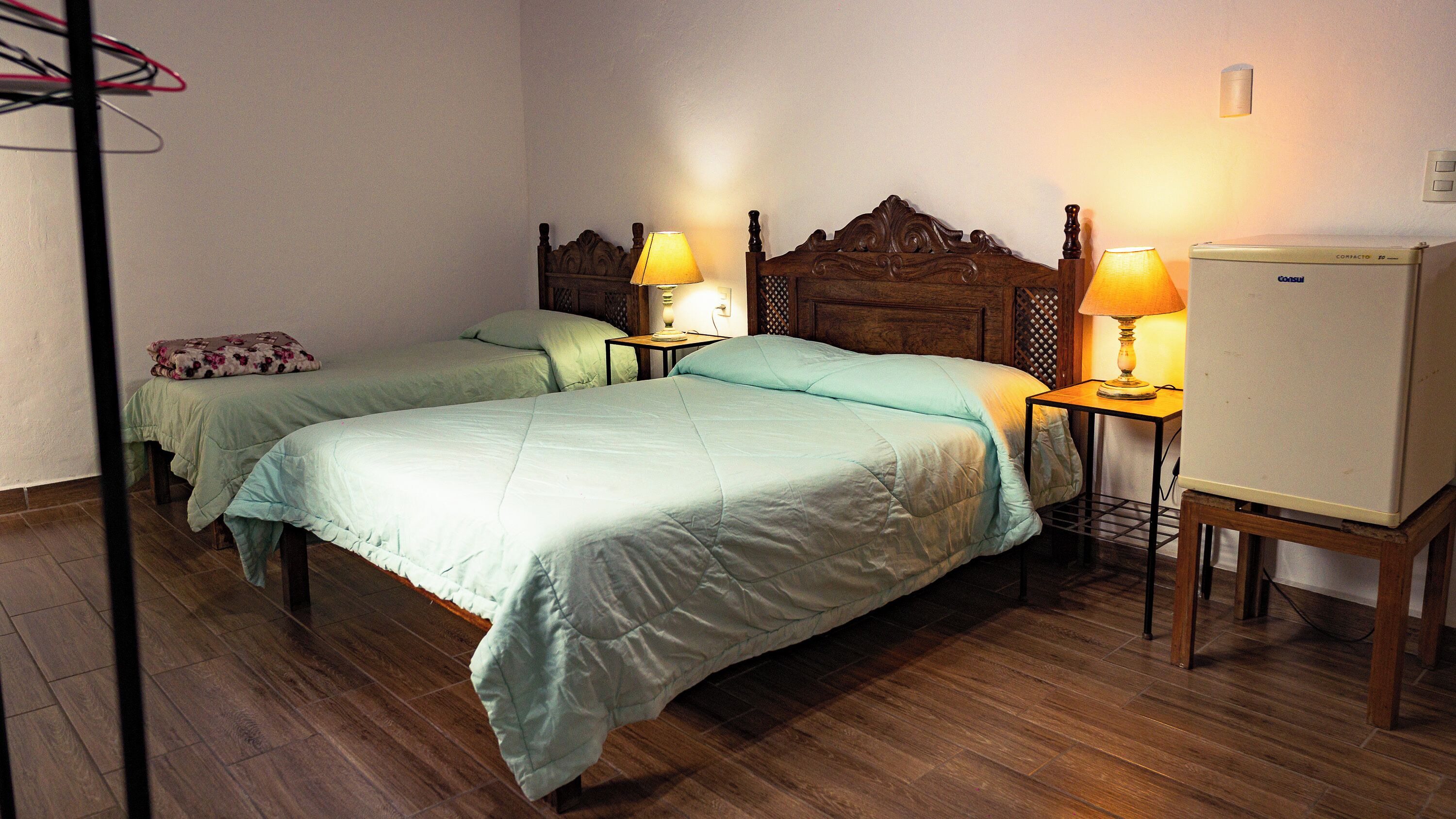 A hotel room with two beds, wooden headboards, bedside lamps, a wooden floor, and a refrigerator.
