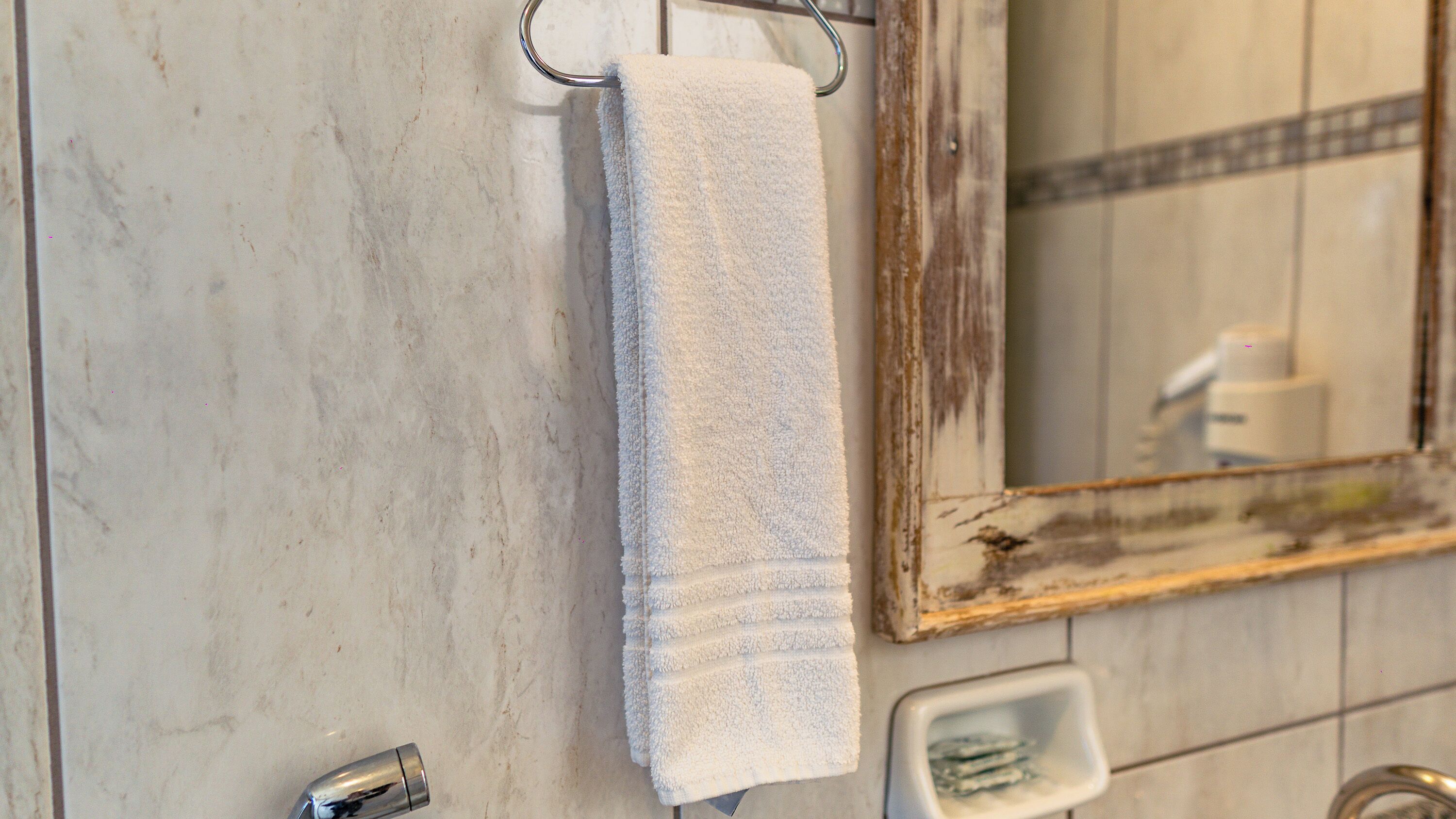 A bathroom with a white towel hanging on a metal towel rack, a mirror with a wooden frame, and a small white soap dish on the tiled floor.