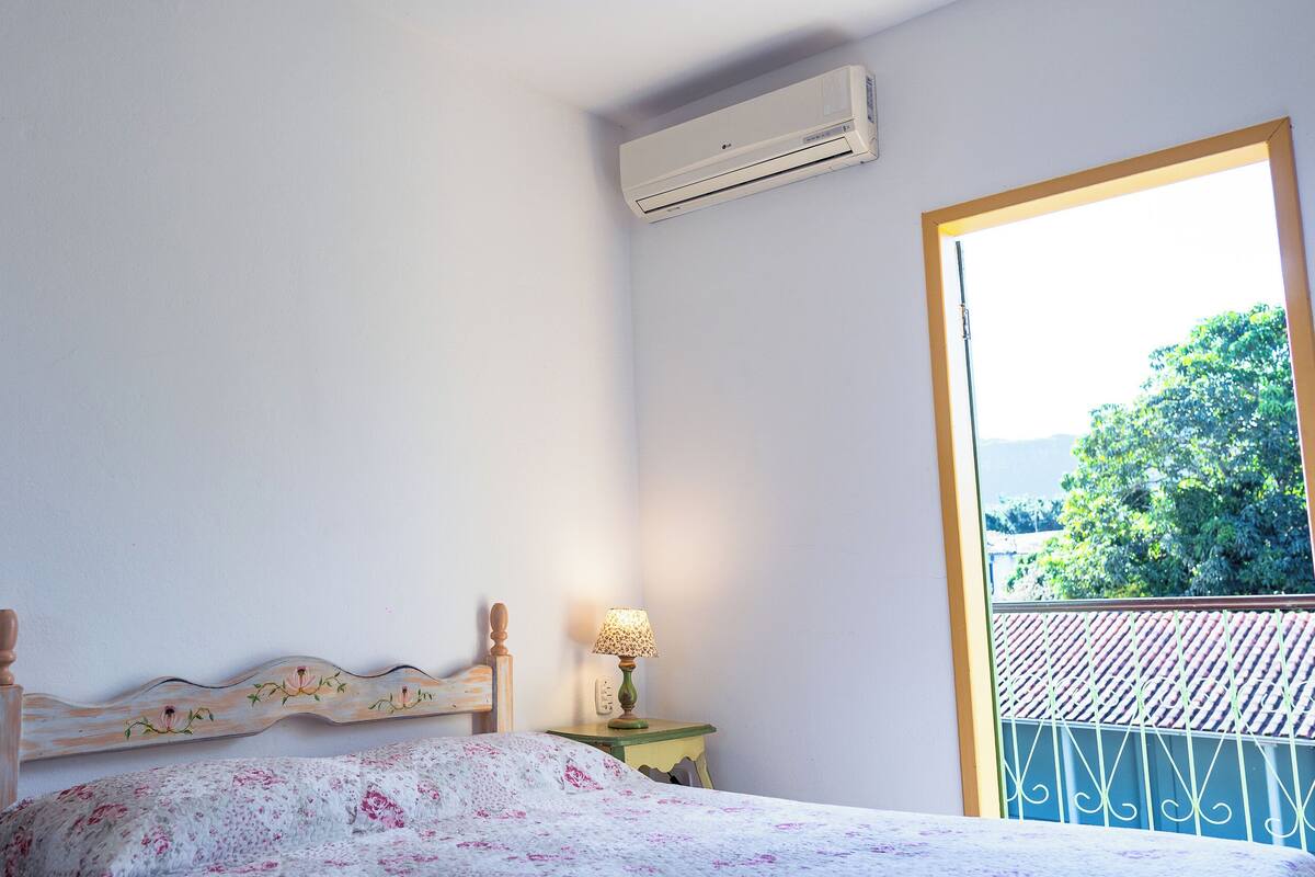 Ein Schlafzimmer mit Bett, Nachttisch, Lampe, Klimaanlage und einem Balkon mit Blick auf Grünflächen.