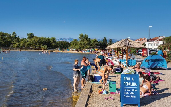 A beach with people sunbathing and swimming, a sign for renting pedalina, and a clear sky.
