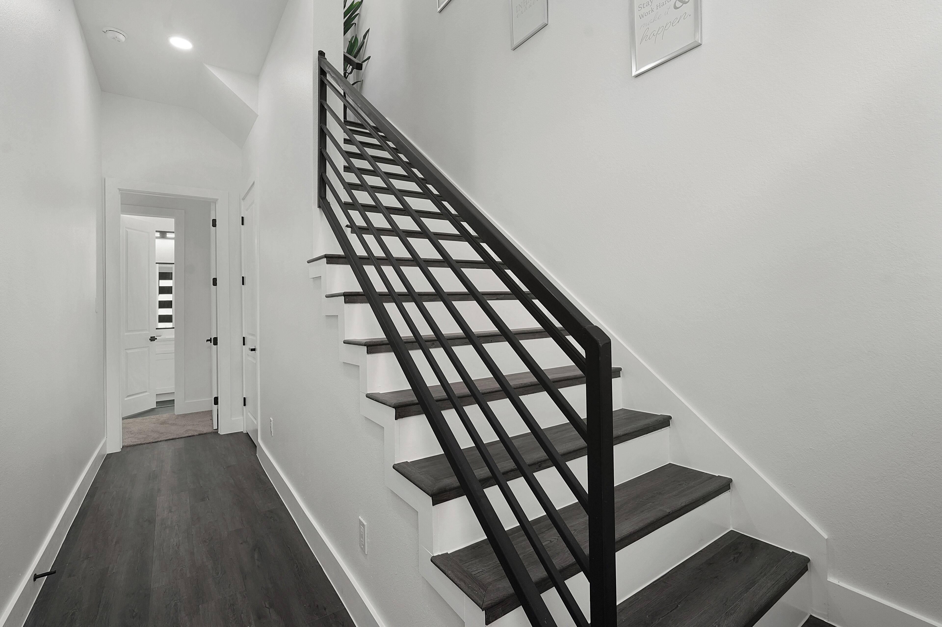 Un escalier avec une rampe en métal noir et des marches en bois, menant à une pièce dotée d’une porte blanche et d’une plante accrochée au mur.