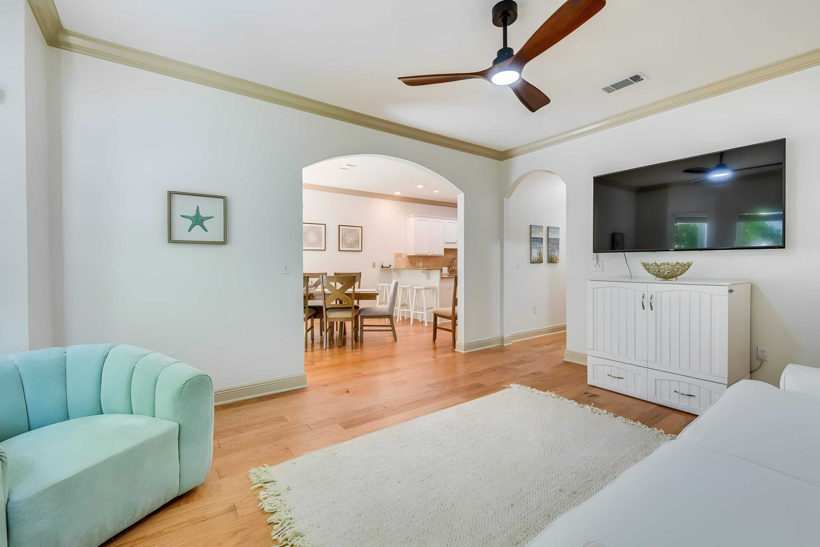 A spacious living room with a ceiling fan, a flat-screen TV mounted on the wall, and a dining area visible through an archway.