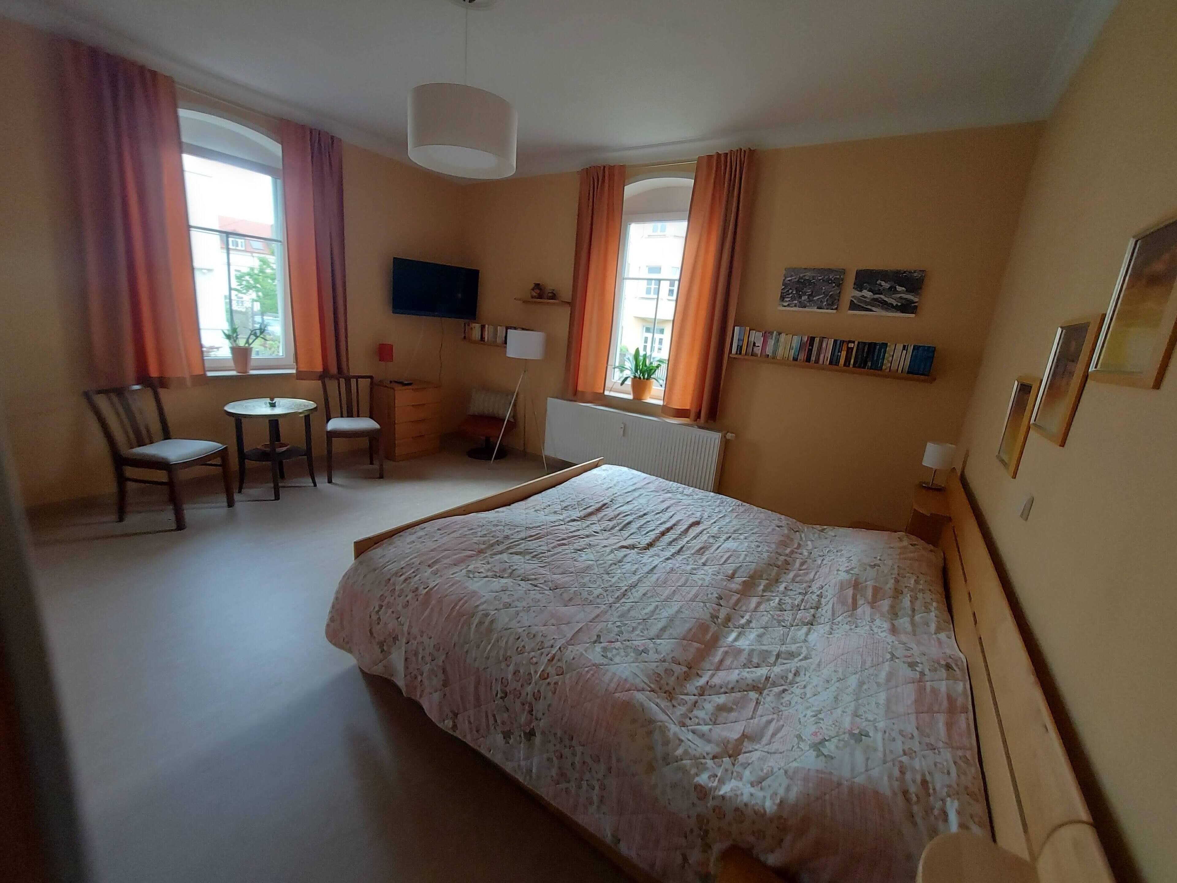 Ein Schlafzimmer mit Bett, Schreibtisch, Stuhl, Fernseher und einem Fenster mit Vorhängen.
