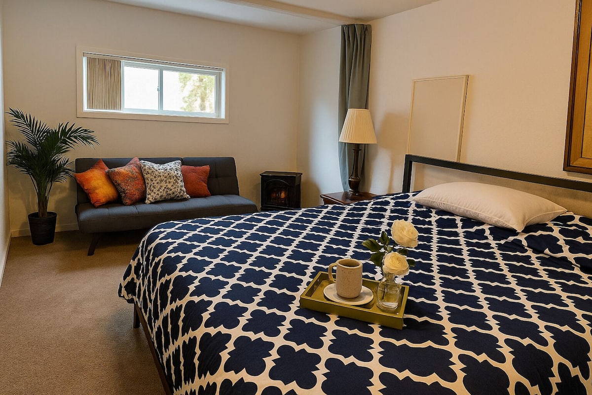 A hotel room with a large bed, a sofa, a potted plant, and a window with blinds.