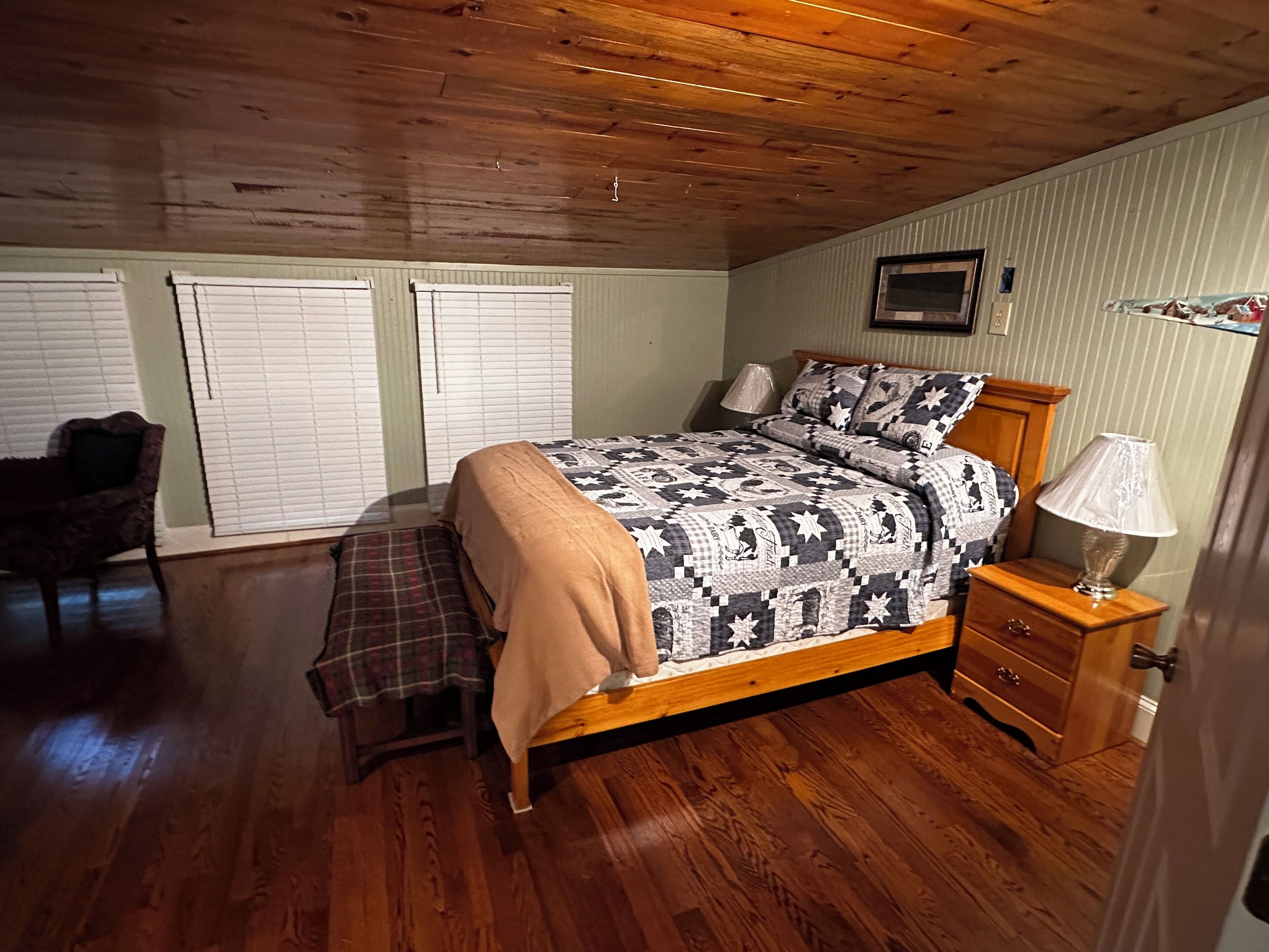 A bedroom with a wooden ceiling, a bed with a patterned quilt, a bench, a nightstand with a lamp, and a chair.