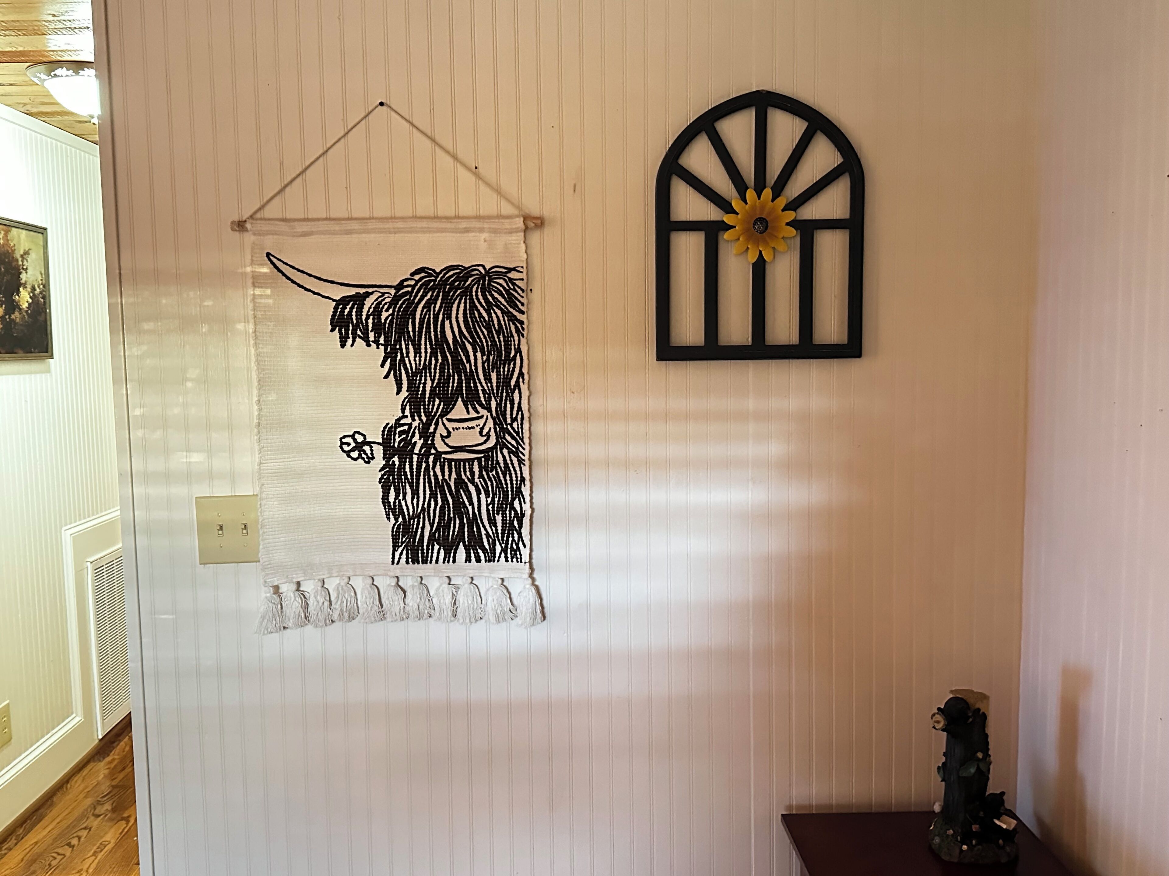 Una pared con una obra de arte en blanco y negro de una vaca, una ventana con marco negro y un girasol, y una pequeña estatua sobre una mesa.