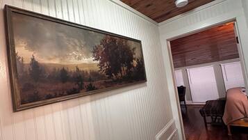 Habitació amb un gran quadre de paisatge a la paret, sostre de fusta i vista cap a una altra habitació amb una finestra i una cadira.