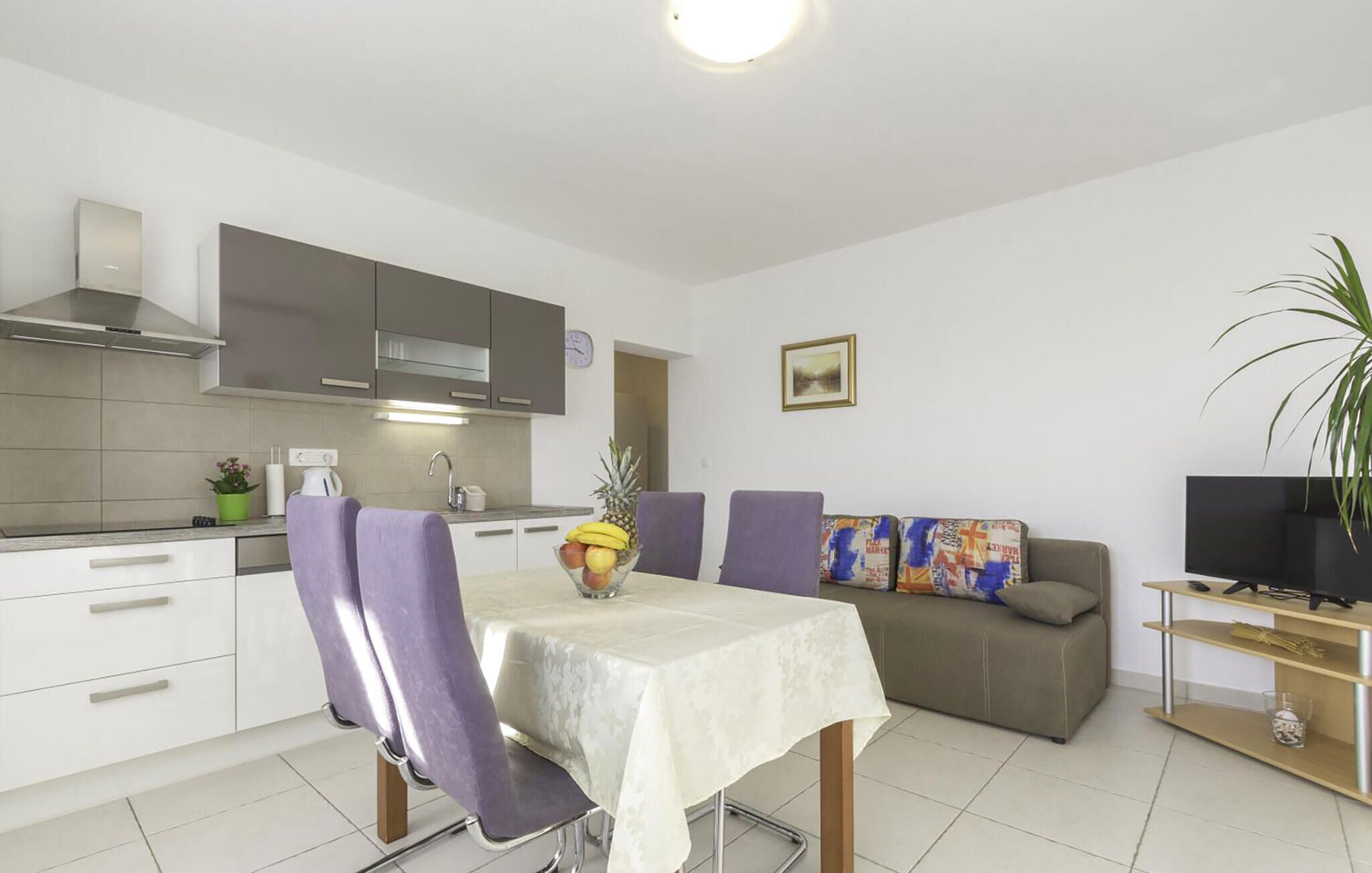 A modern kitchen with white cabinets, a dining table with purple chairs, a sofa with colorful cushions, and a TV on a wooden stand.