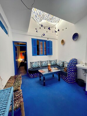 Une pièce avec un tapis bleu, un canapé orné de coussins à motifs, une table basse et une lucarne décorée de suspensions.