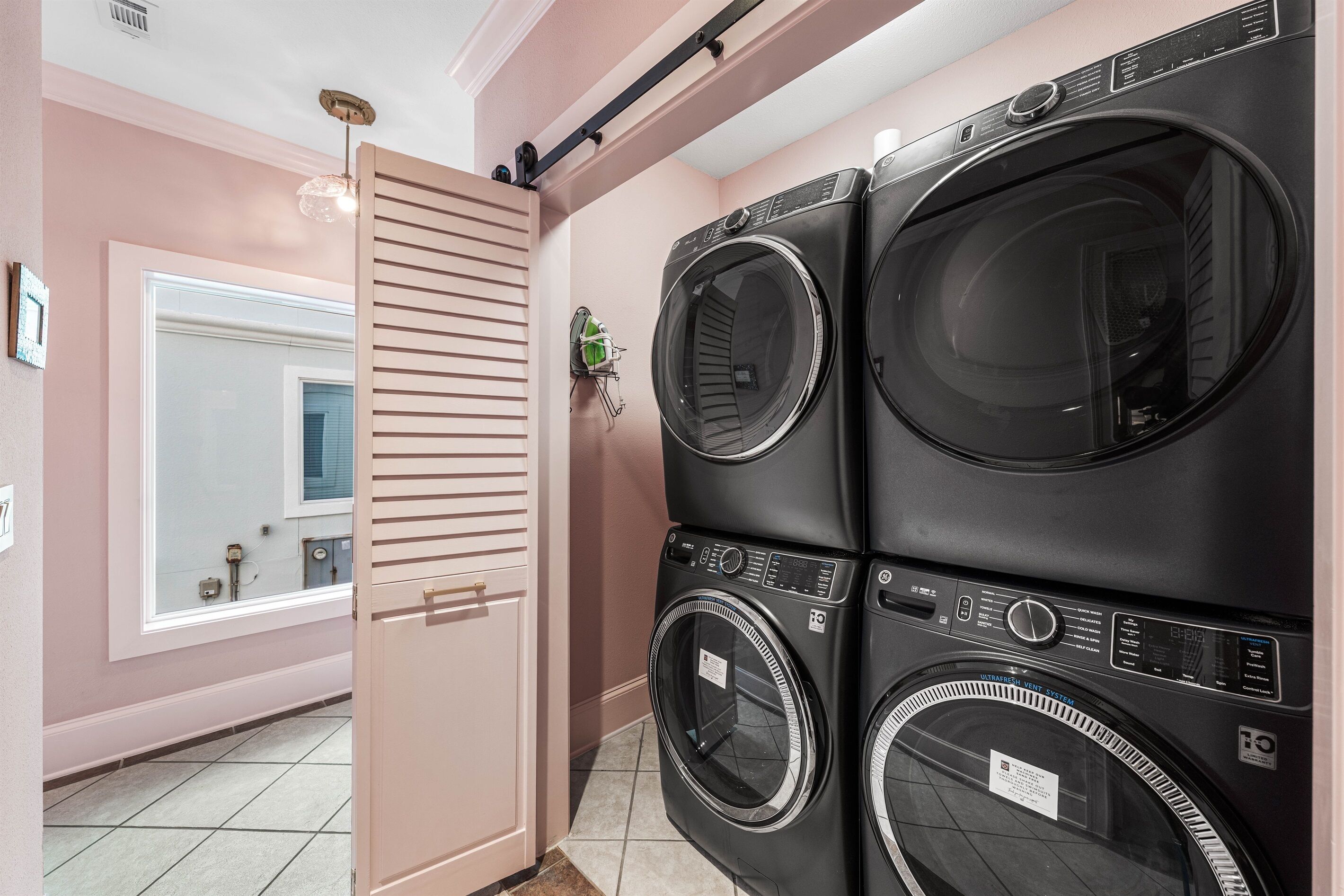 A laundry room with two stacked front-loading washers and dryers, a sliding door, and a window with blinds.