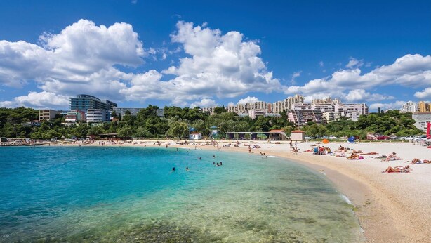 A beach with clear turquoise water, a sandy shore, and a backdrop of multi-story buildings. - Gorgeous home in Mravince (Mravince)