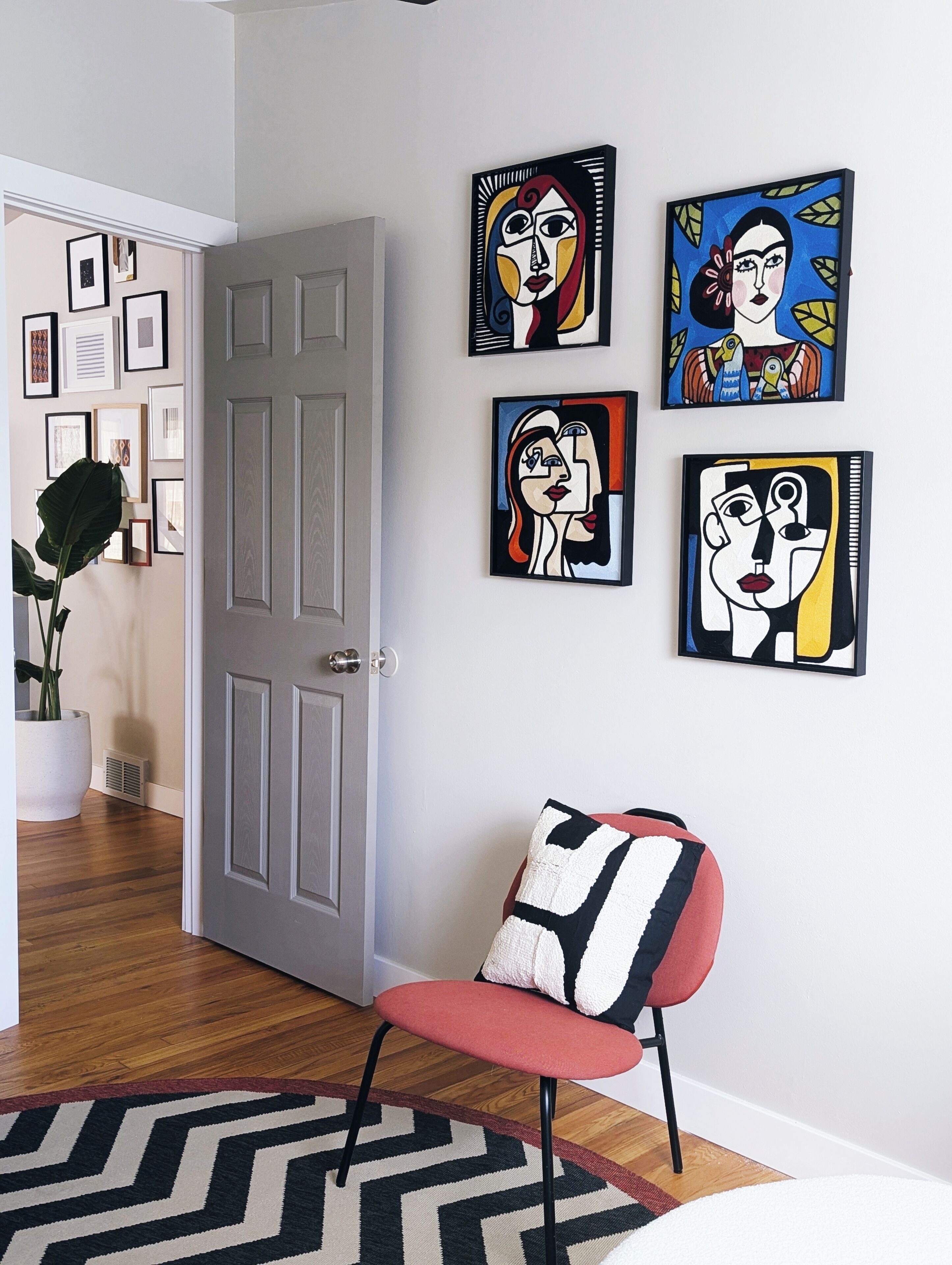 A room with a grey door, a red chair with a black and white pillow, and four abstract paintings on the wall.