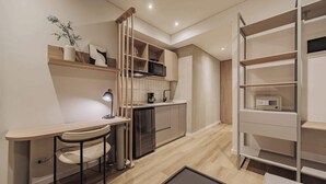 Una cocina compacta con microondas, cafetera y un mueble pequeño. Hay un escritorio con silla y una lámpara, además de una estantería con algunos objetos.