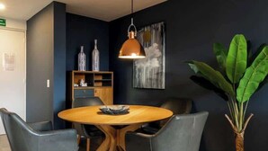 Un coin repas avec une table ronde en bois, quatre chaises grises, une suspension en cuivre, une bibliothèque agrémentée d’objets décoratifs et une grande plante verte.