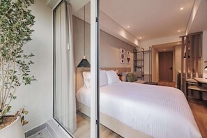 A modern bedroom with a large bed, a bedside table, a lamp, and a potted plant. - Quadrato | Laureles Studio, w/AC, Balcony, WIFI (Medellín)