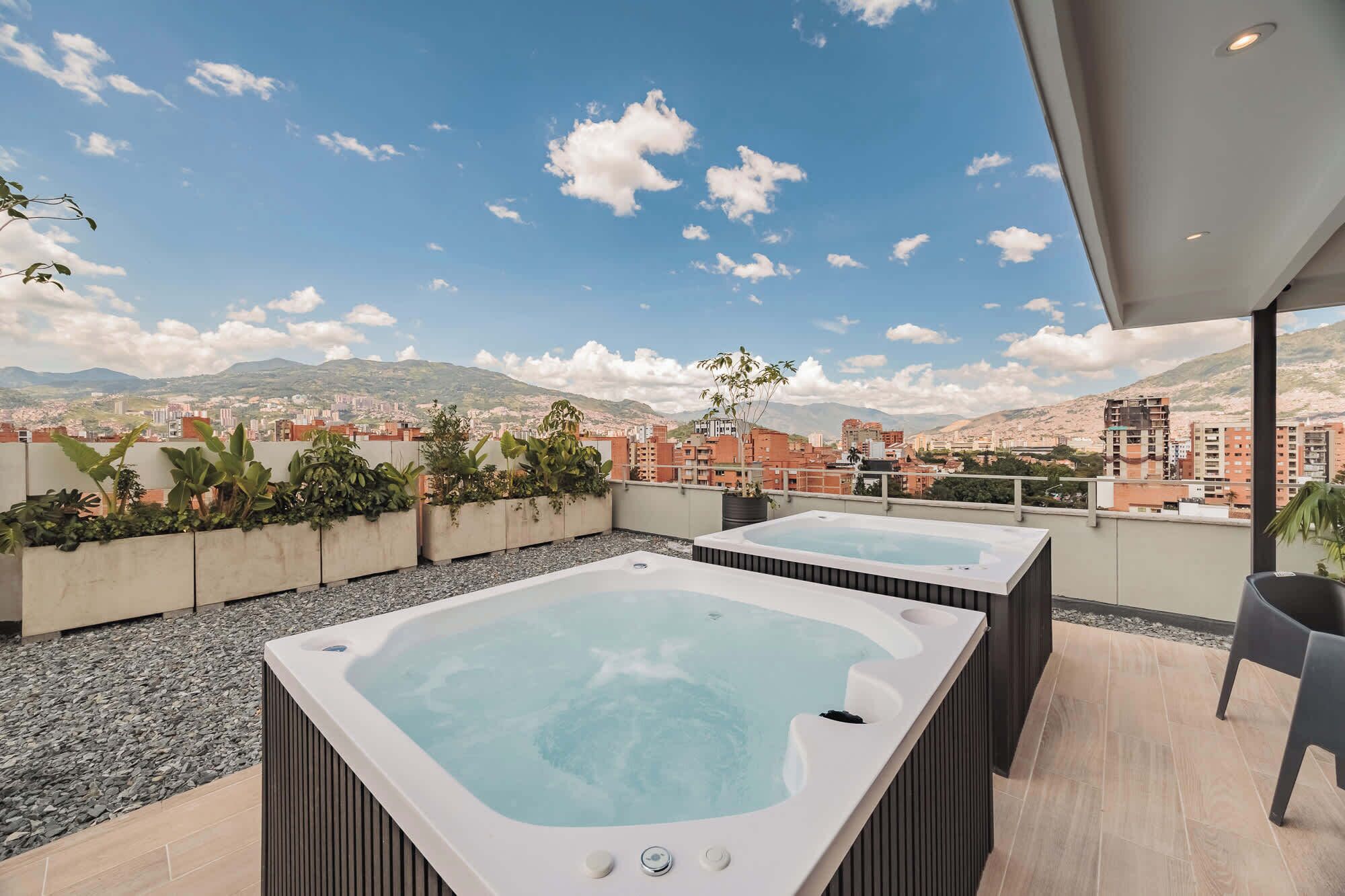 A rooftop terrace with two hot tubs, a seating area, and a view of a cityscape with mountains in the background.