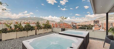 A rooftop terrace with two hot tubs, a seating area, and a view of a cityscape with mountains in the background.