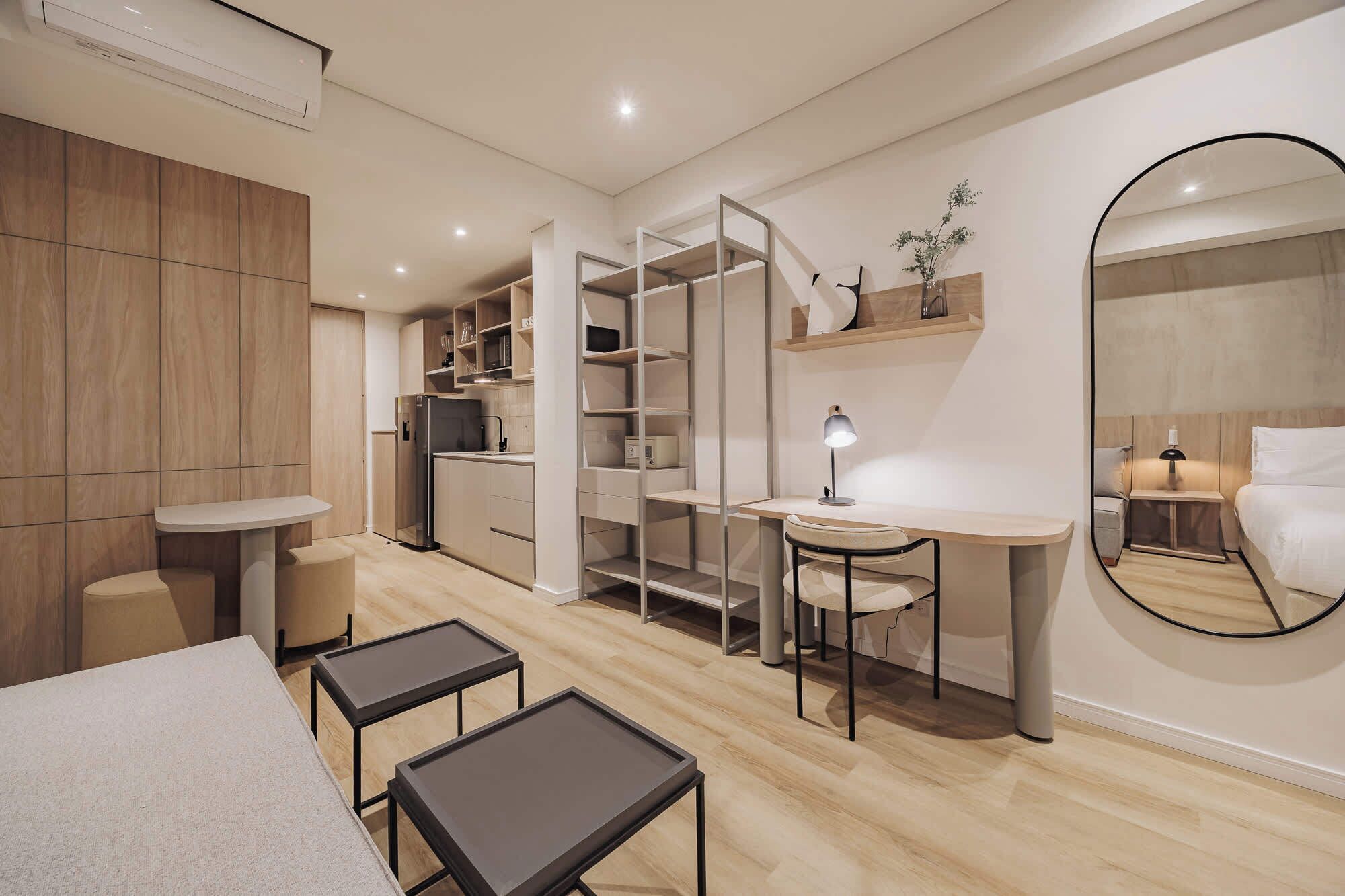 Un dormitorio moderno y compacto con cama, escritorio y silla. Hay un armario con puertas abiertas, un espejo y una mesita de noche con lámpara.