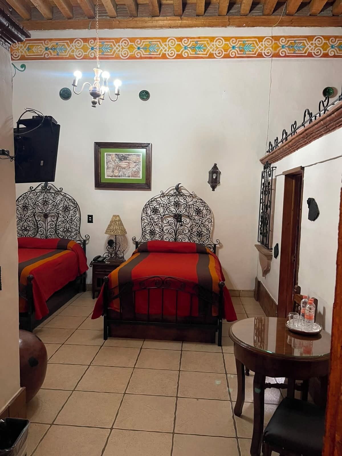 A room with two beds, a chandelier, a framed picture, a small table with glasses, and a door with a decorative metal railing.
