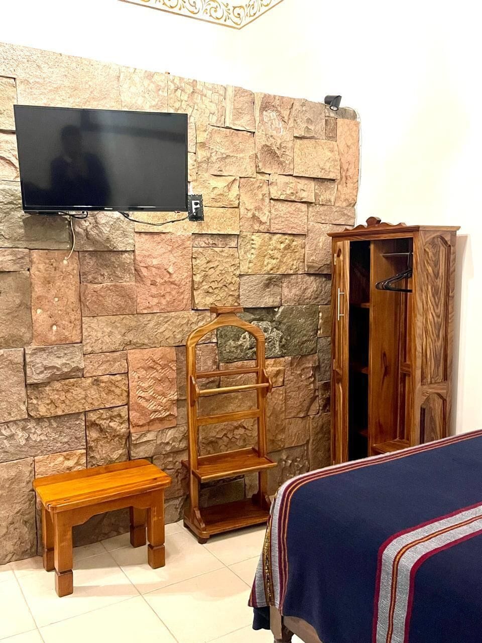 Una habitación con pared de piedra, un televisor de pantalla plana montado sobre un banco de madera, un armario de madera y una cama con colcha a rayas.