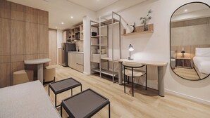 A modern, compact bedroom with a bed, a desk, and a chair. There is a wardrobe with open doors, a mirror, and a small table with a lamp. - Quadrato | Laureles Studio, w/AC, Balcony, WIFI (Medellín)