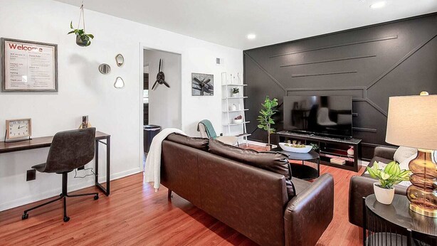Una sala de estar moderna con un sofá de cuero marrón, un televisor de pantalla plana y piso de madera. Hay un escritorio con una silla y una planta pequeña en la pared.