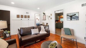 Una sala de estar moderna con un sofá de cuero marrón, una mesita de centro redonda y un sillón beige. Se ve una cocina al fondo con una mesa de comedor y sillas.