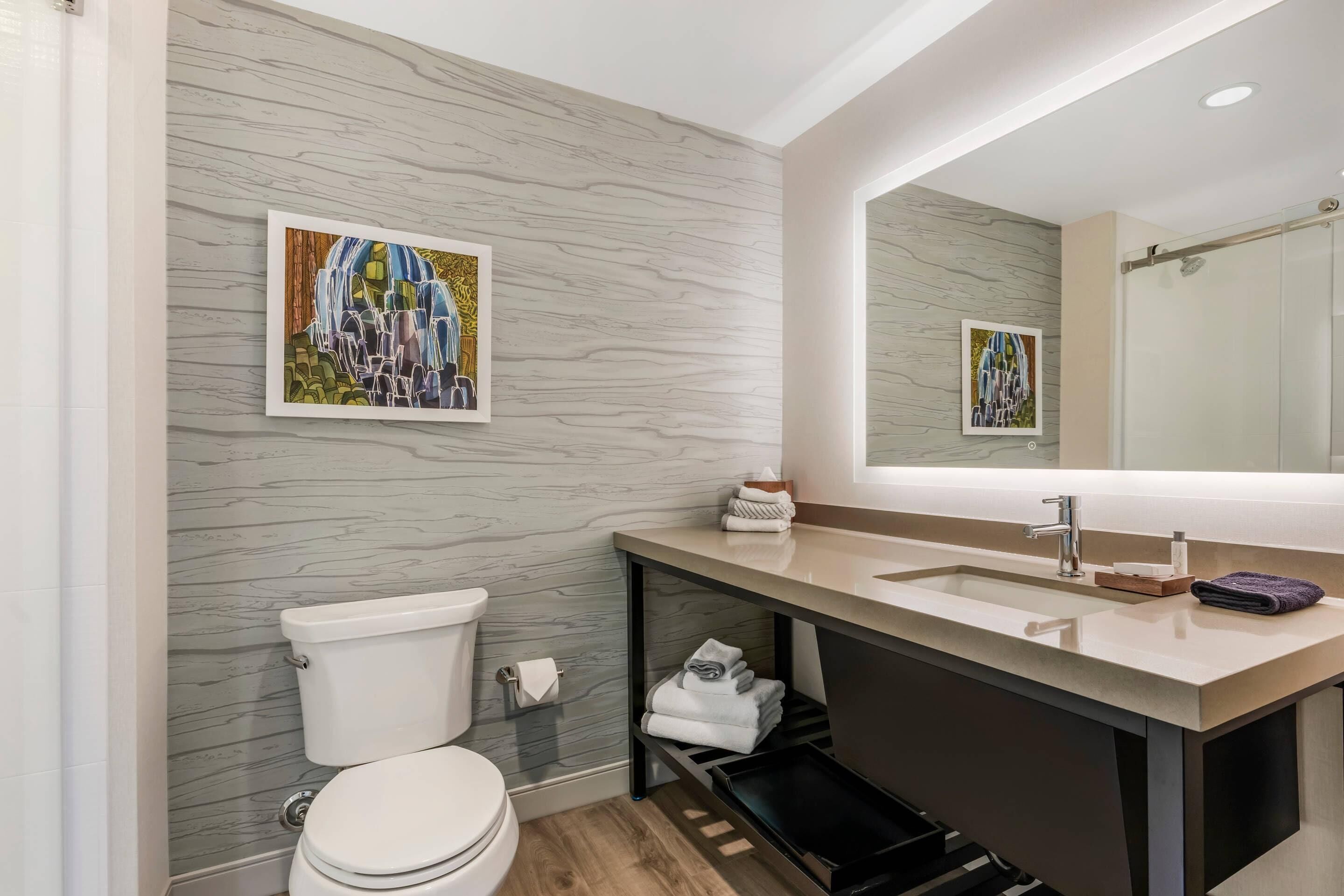 A modern bathroom with a marble wall, a toilet, a sink with a mirror, and two framed artworks.