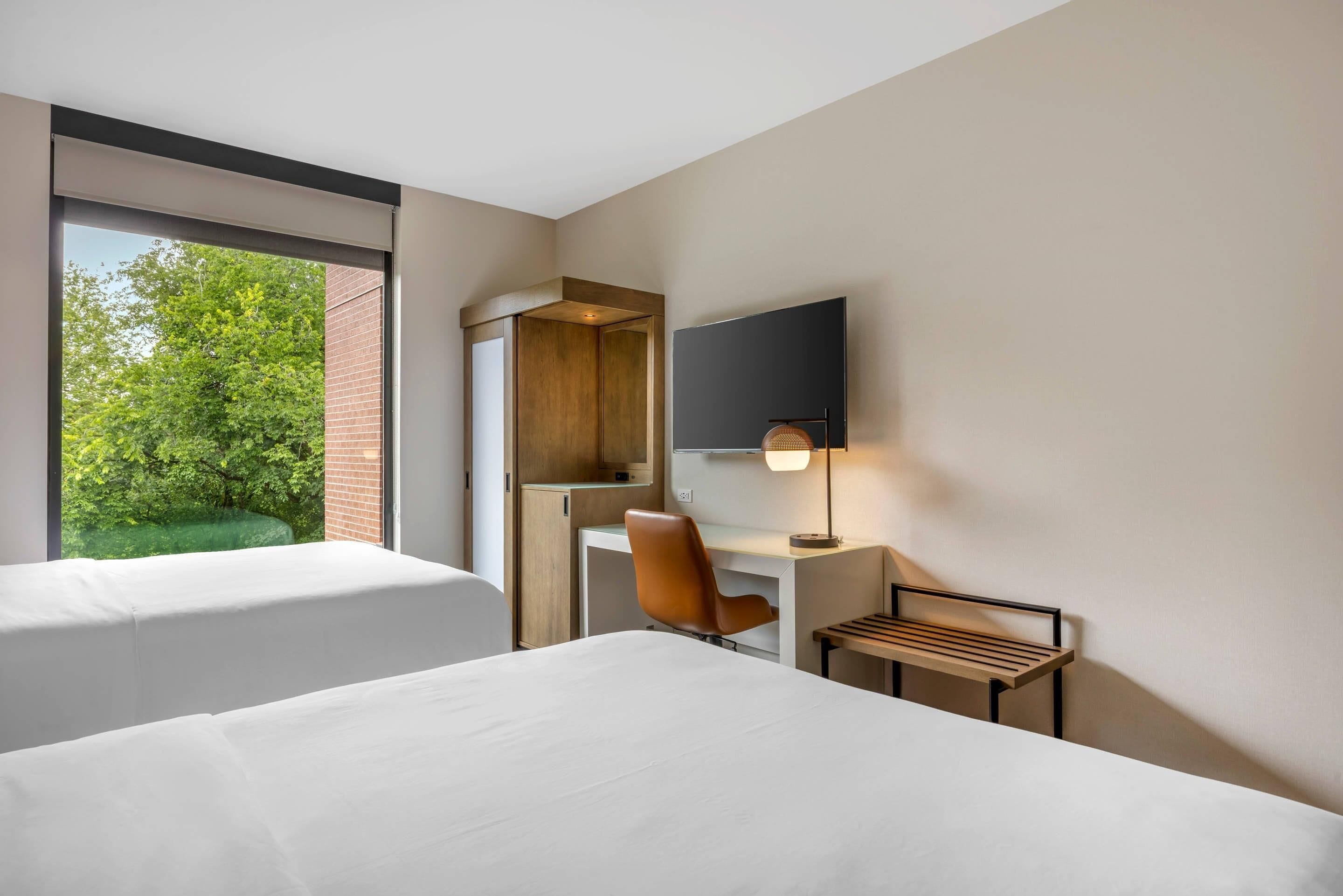 A hotel room with two beds, a desk, a chair, a TV, and a window with a view of greenery.