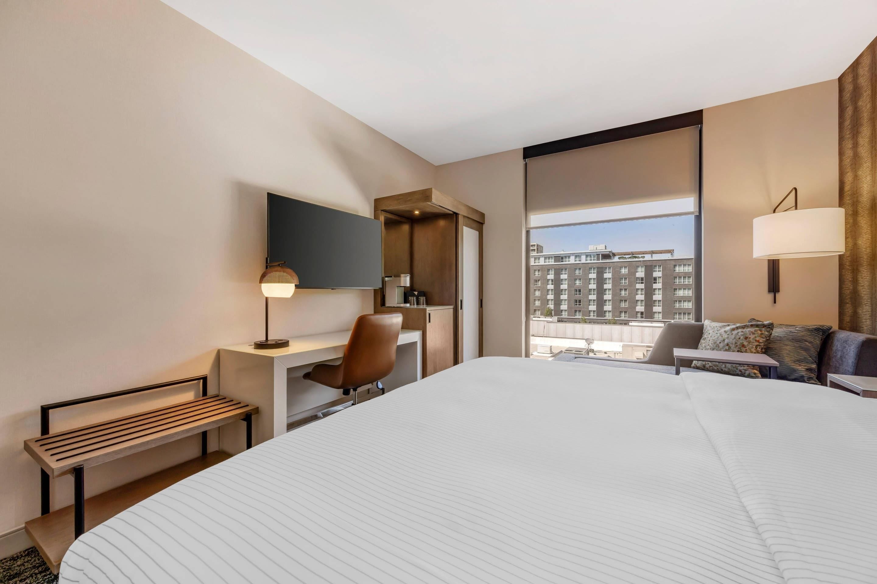 Habitación de hotel con una cama grande, un escritorio con silla, un televisor montado en la pared, una ventana con vista a edificios y una lámpara en la mesita de noche.