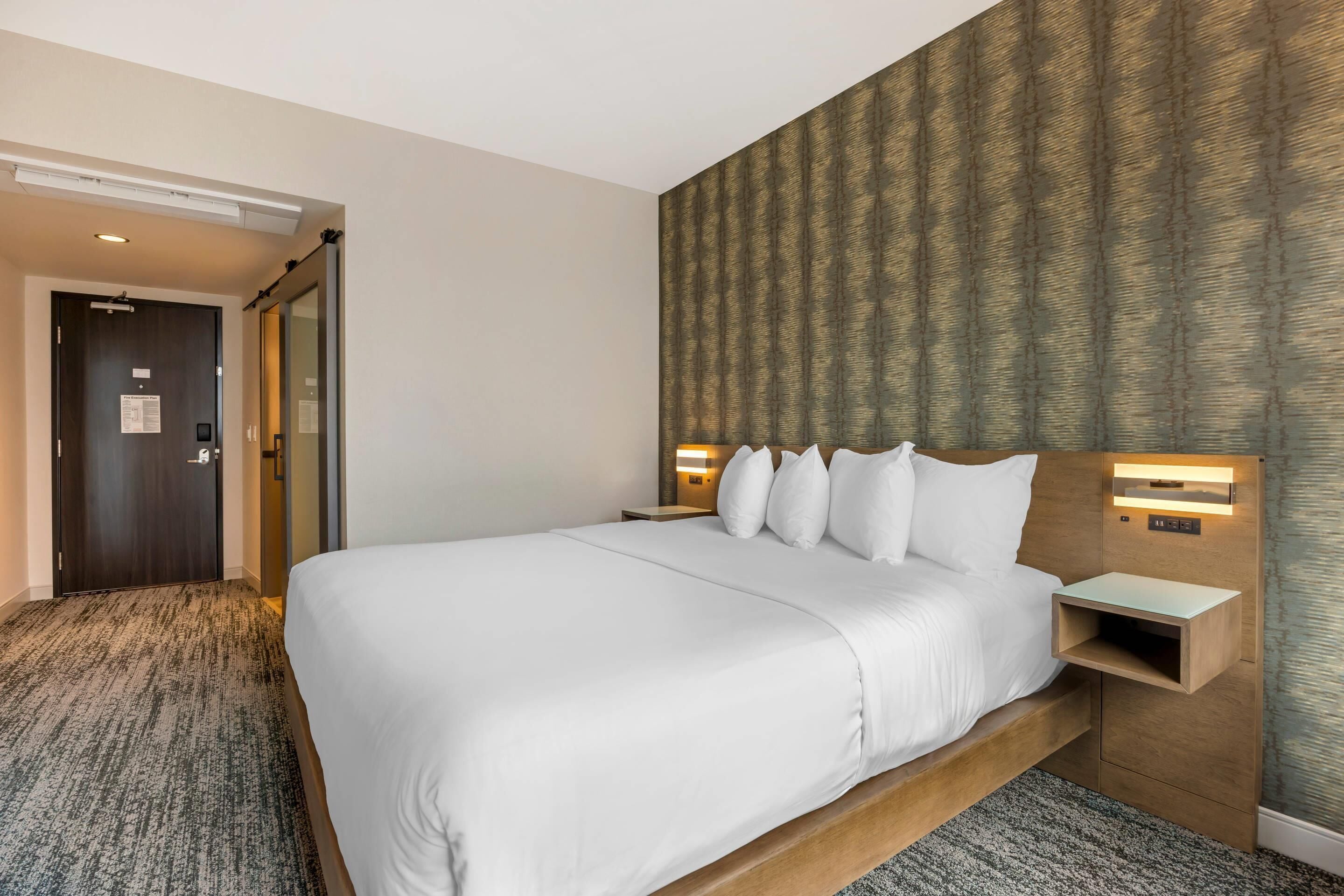 A hotel room with a large bed, a wooden headboard, two bedside tables with lamps, a carpeted floor, and a patterned wallpaper on the wall.