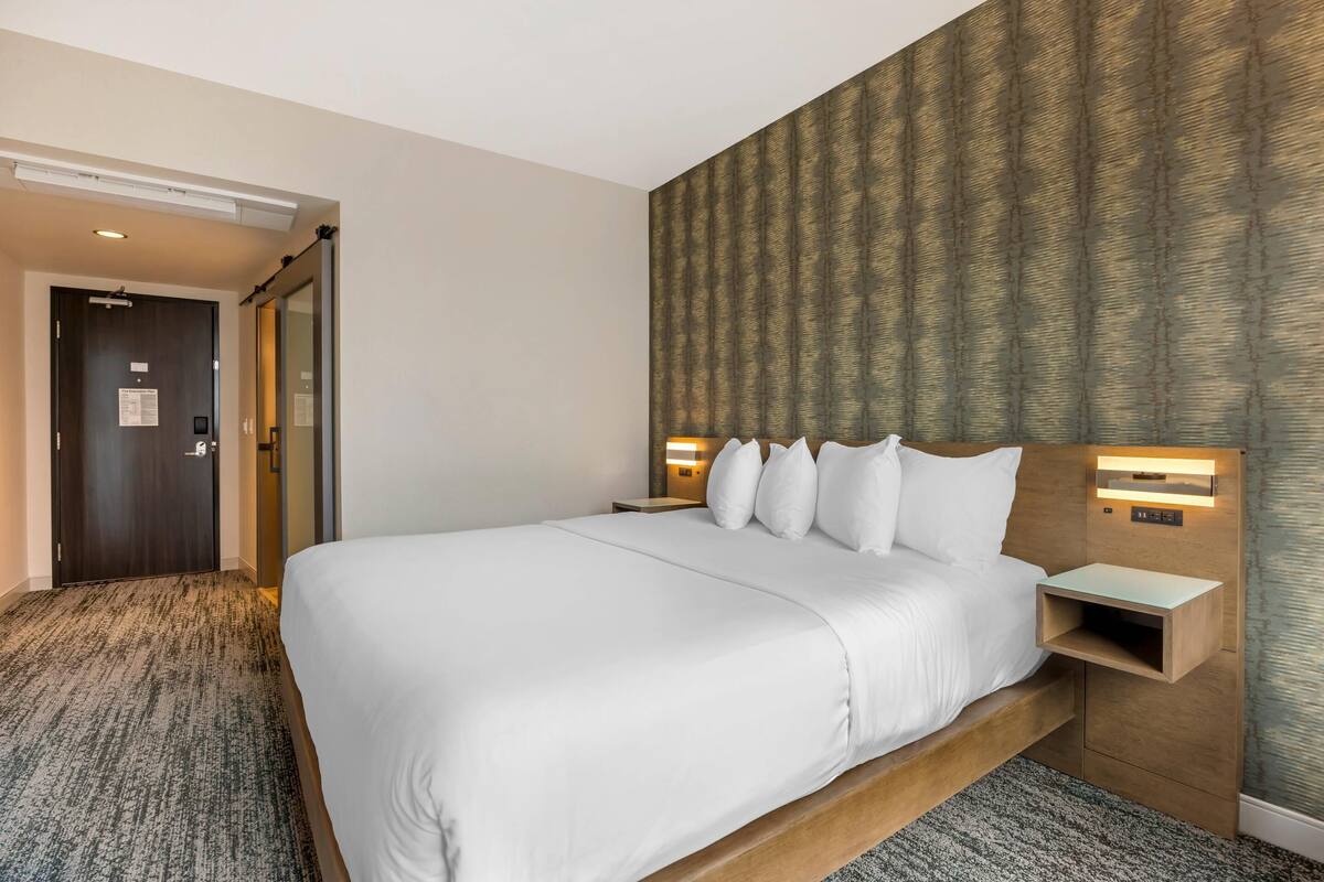 Une chambre d’hôtel avec un grand lit, une tête de lit en bois, deux tables de chevet avec des lampes, un sol recouvert de moquette et un papier peint à motifs sur le mur.