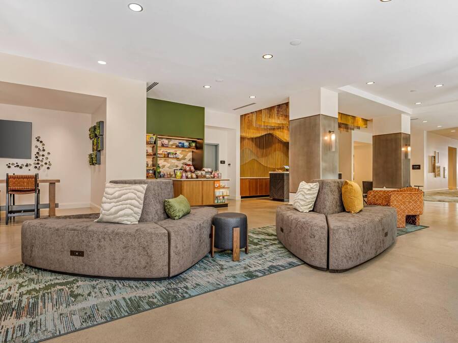 A spacious living area with two grey sofas, a coffee table, and a TV mounted on the wall. There is a kitchen area in the background with a wooden cabinet and a refrigerator.